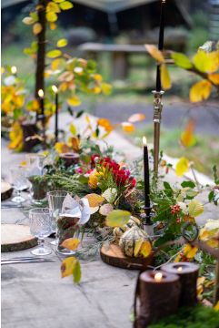 An autumn colored table arrengement that includes red flowers, yellow leafs, brown candles and Nomadic's unique wood plates