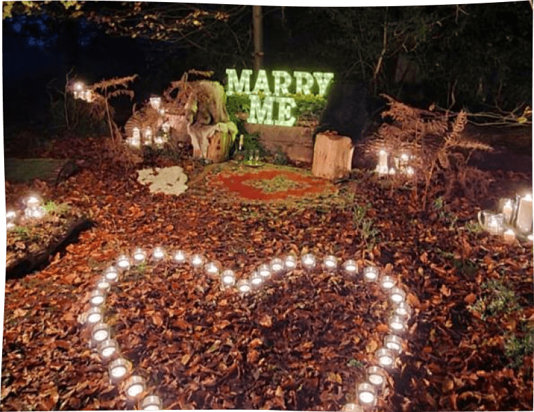 Corporeal "marry me" lighting letters, next to a heart-shape candle arrengement for a woodland proposal